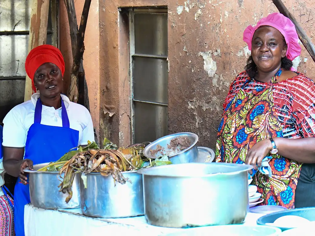 Two cooks in bright clothes serving food from large pots